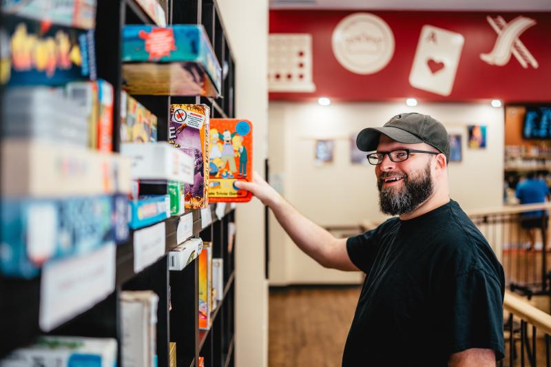 A bearded man wearing a black t-shirt, glasses, and a baseball cap smiles while reaching for a board game on a well-stocked shelf in a board game café.