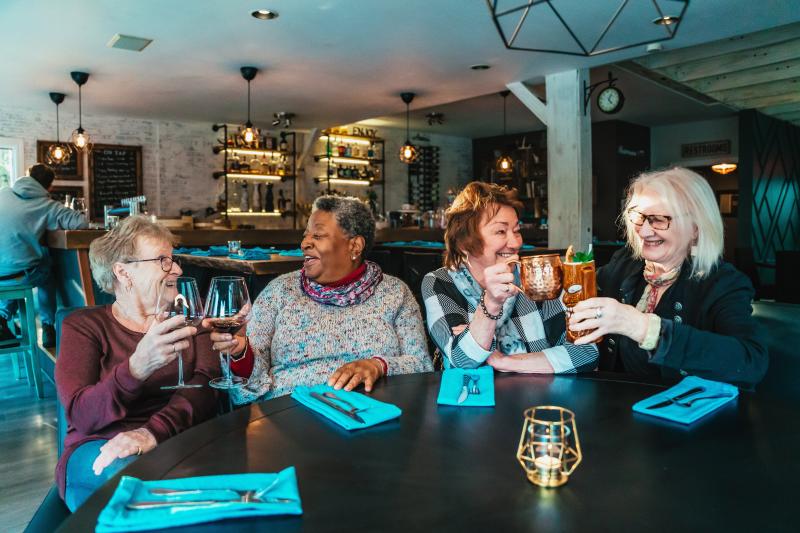 Four older women sit around a table in a cozy restaurant, smiling and raising glasses in a toast. Two hold red wine while the others hold copper mugs and cocktails, with warm lighting and a rustic bar visible in the background.