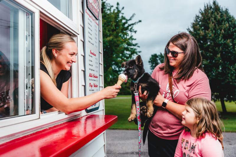 A smiling ice cream stand employee hands a small vanilla cone to a black and tan puppy being held by a woman outdoors, while a young child looks on excitedly.