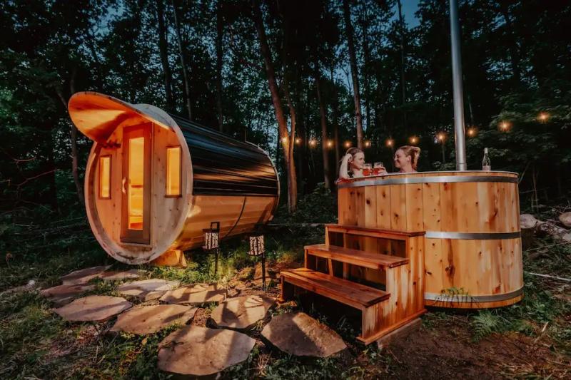 Two people relax in a round wooden hot tub at Oliver’s Tiny Home in the Forest. It's dusk, surrounded by trees and string lights. Next to them is a barrel-shaped wooden sauna with a softly glowing interior light. A stone path leads to the sauna, and the cozy scene is set in a peaceful forest setting.