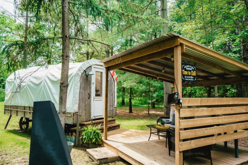 A covered wagon-style glamping cabin with a wooden deck and covered seating area surrounded by tall trees in a forest setting.