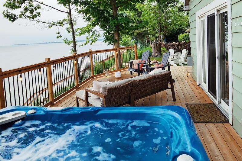 A scenic lakeside deck in Selkirk Ontatrio featuring a bubbling blue hot tub in the foreground. The wooden deck is furnished with cushioned seating, Adirondack chairs, and a round table set with wine glasses, surrounded by string lights along the railing. Lush green trees frame the view, and the calm lake stretches out in the background under a soft, overcast sky. A sliding glass door leads inside the house on the right.