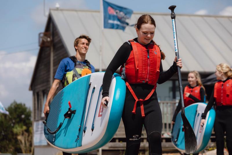 A woman wearing a red life vest carrying a blue paddleboard with her instructor, at Eastbourne