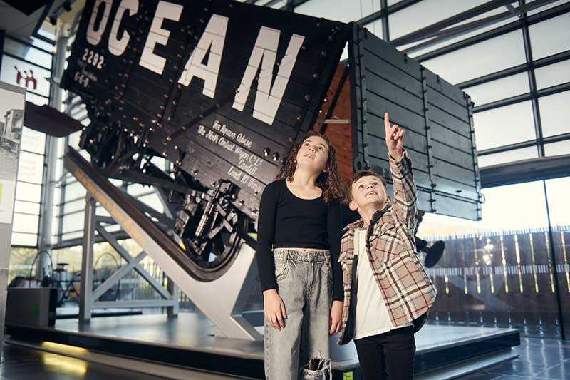 Two children standing in front of an old railway cart in the National Waterfront Museum.