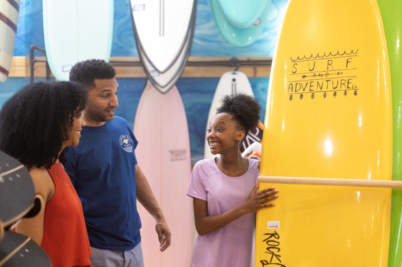 a man and woman talking to a girl propping up a yellow surfboard talking inside of a surf shop