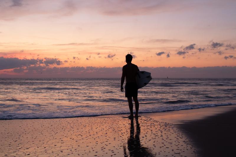 silhouette of a man holding a surfboard standing at the water's edge at sunrise