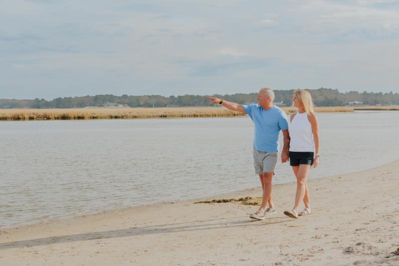 man wearing a blue polo and khaki shorts holding woman wearing white tank top and black shorts hands walking across shoreline and pointing to something in the distance