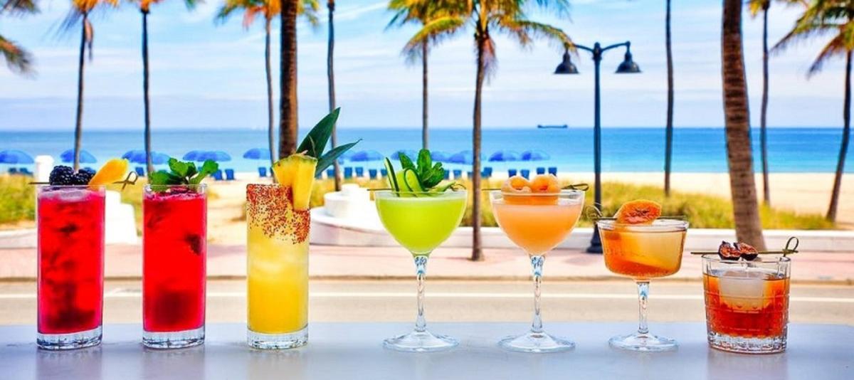 A selection of 7 craft cocktails on a counter with the ocean in the background