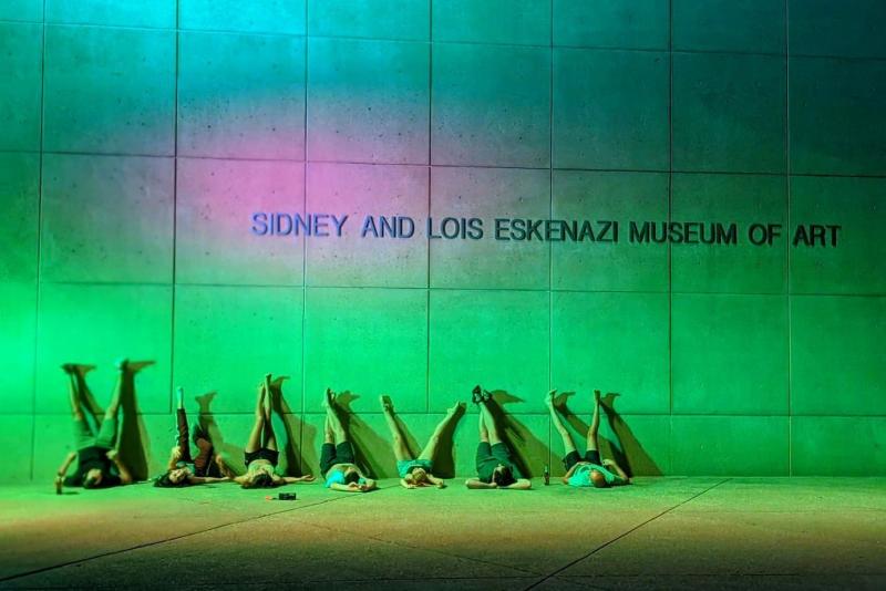 Seven people laying on the ground with their legs propped against the Eskenazi Museum of Art watching the light totem change colors