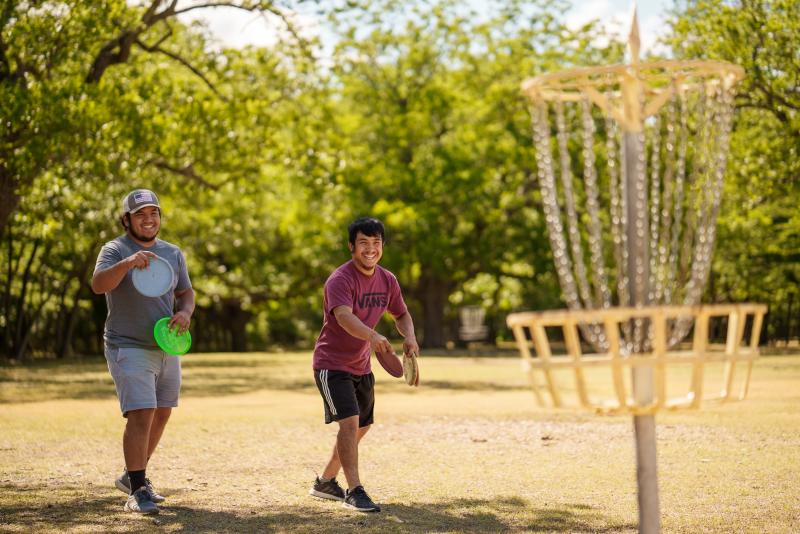 Two men smiling, playing disc golf, throwing discs into a basket at Riverside Park.