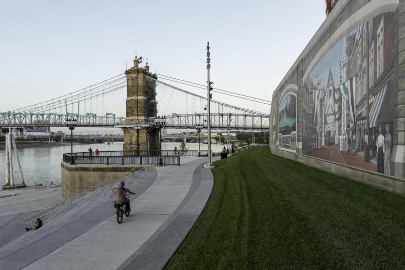 Along the right side are some of the murals from the Roebling Mural walk. In the foreground people walk, bike, sit, and watch the river. The bridge is in the background.