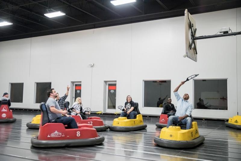 A group of adults riding on bumper cars reaching for a whiffleball at WhirlyBall