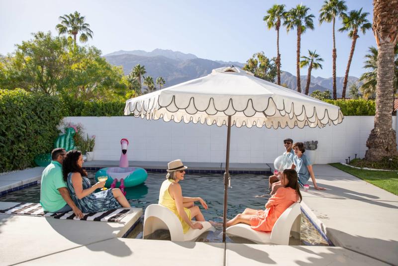 Group of people enjoying a sunny day by an outdoor pool with a mountain backdrop, with lounge chairs, a pool umbrella, and inflatable pool toys visible.