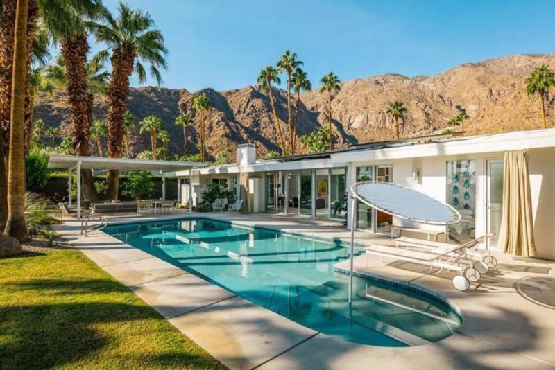 An image of a luxury mid-century modern home with a swimming pool in the foreground, surrounded by palm trees, with a desert mountain landscape in the background. The house features large windows, white exterior walls, and a flat roof, with outdoor furniture and sun loungers arranged on the poolside patio.