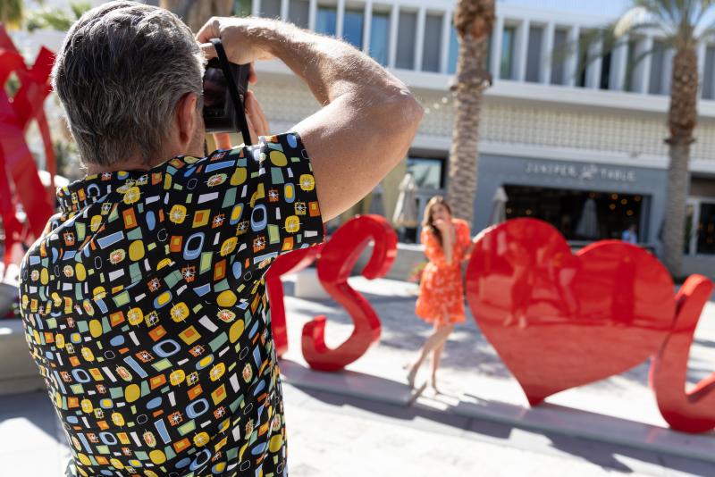 Man in patterned shirt taking a photograph of a woman in an orange dress posing among large red heart sculptures outdoors.