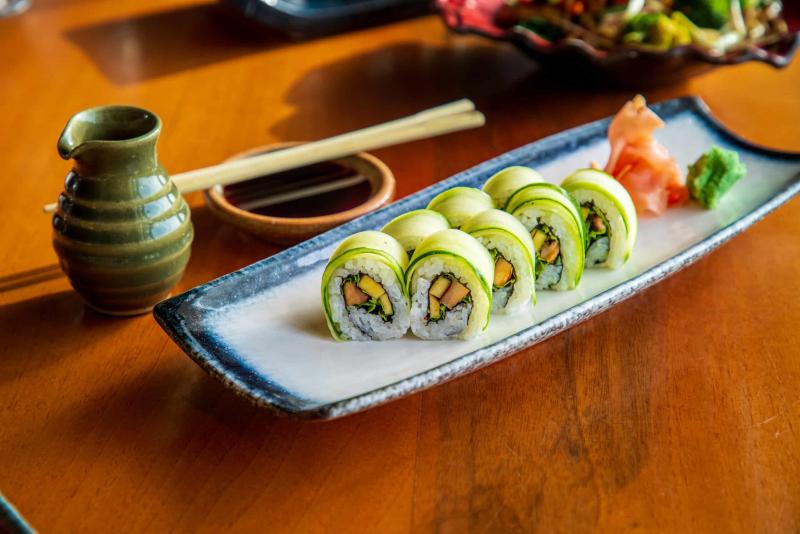 Cucumber coated sushi rolls served with ginger and wasabi on rectangular plate, soy sauce with chopsticks on the table