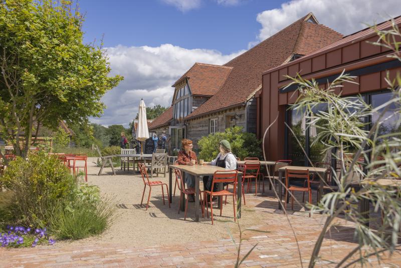 A couple sit at a table in a sunny courtyard surrounded by plants, trees and the Knepp kitchen buildings