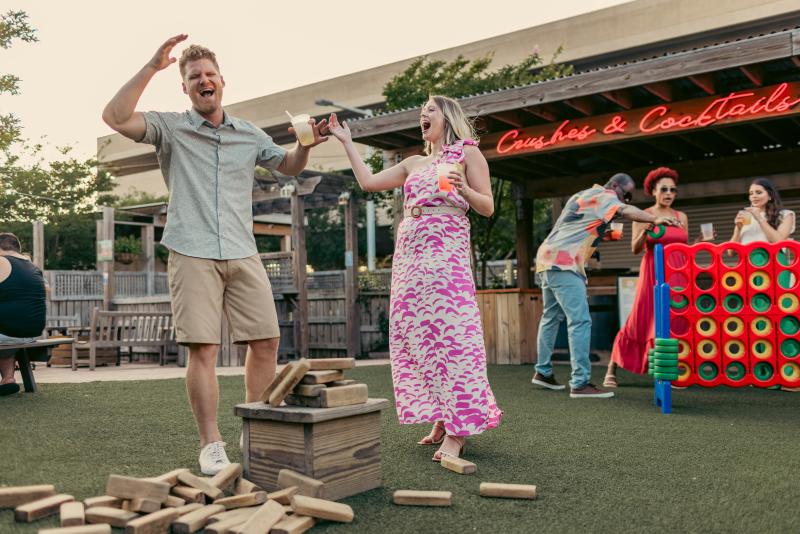 Two adults holding cocktails playing giant jenga blocks and connect four at oceanfront bar