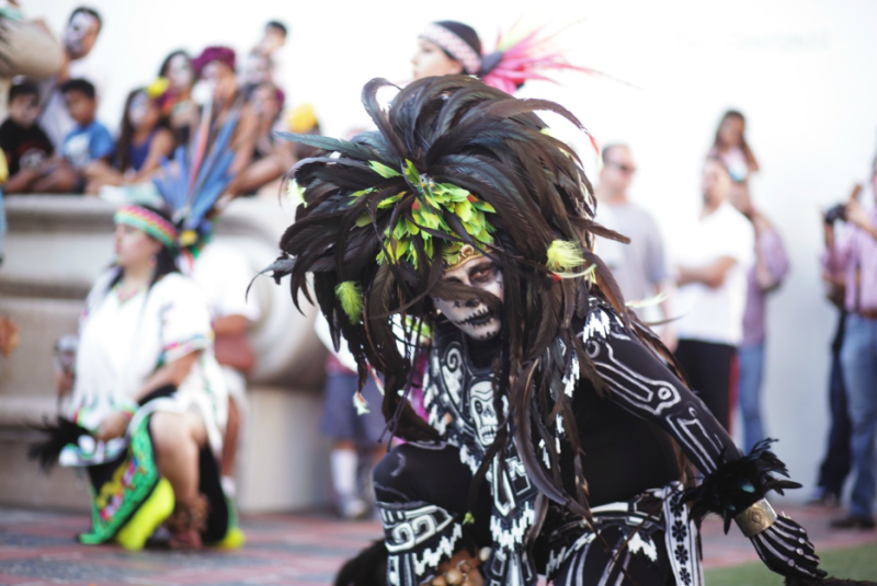 A Día de los Muertos dancer in traditional Aztec attire with a Catrina-style face painting, performing at a public celebration. The dancer wears a vibrant feathered headdress and skeletal body paint, honoring Mexican culture and traditions.