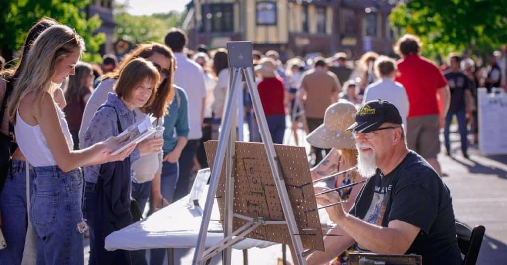 Person painting on a canvas in The Village of Wauwatosa, WA. They are surrounded by people watching them paint.