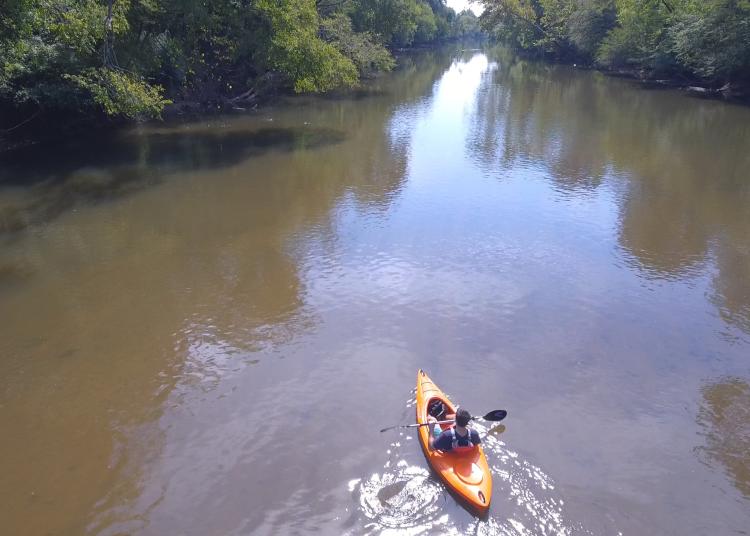 A kayaker paddles a river in the sun captured by a drone.