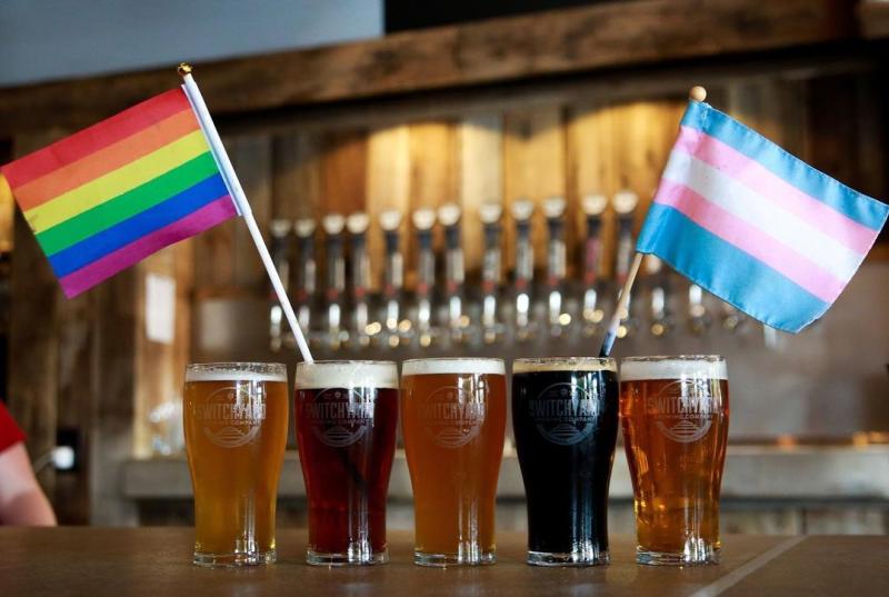 Five beers lined up with the Pride and Transgender flags for Pride Month