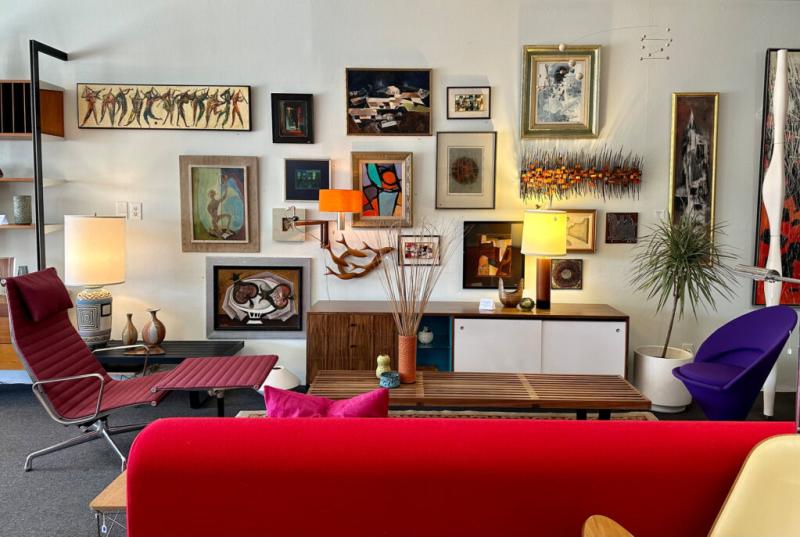 An eclectic art gallery interior with various framed paintings adorning the walls, modernist-style furniture including a red chaise longue and a purple chair, floor lamps, decorative vases, and a tall plant pot.