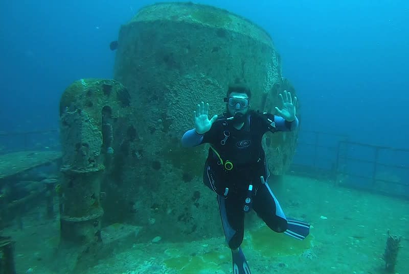 an underwater scuba diver waving at the camera while in front of a section of a shipwreck