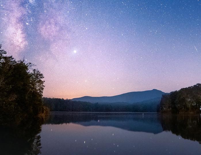 The Milky Way Galaxy can be seen shining above the nighttime silhouette of Grandfather Mountain. The star-filled sky can be seen in the reflection of Julian Price Lake in the foreground.