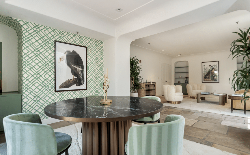 A modern, spacious living room with a black marble round table and green velvet chairs in the foreground. The walls are decorated with geometric-patterned wallpaper and framed bird photographs. The background shows a seating area with cream-colored armchairs, plants, and tasteful decor.