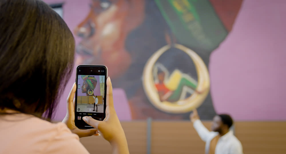 A female taking a photo of a male posing with a mural painted by a Black artist.