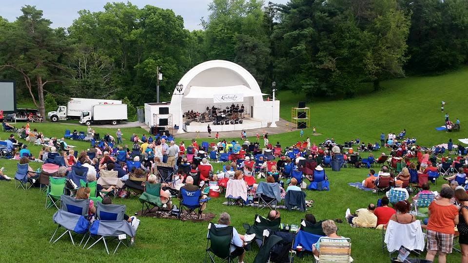 Image is of the outdoor Band Shell amphitheater during a summer day with people sitting in lawn chairs all around it.