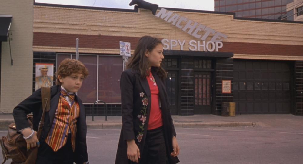 Spy Kids screengrab showing Carmen and Juni walking together in front of a building with sign reading Machete Spy Shop