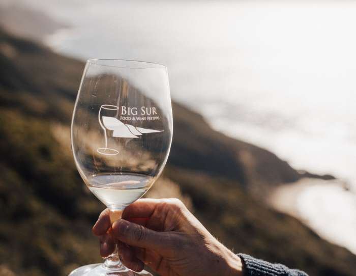 This is an image of a clear commemorative glass from Big Sur Food & Wine Festival held up against the dramatic coastline backdrop of Big Sur, California. The rolling mountains lead straight in to the ocean waters