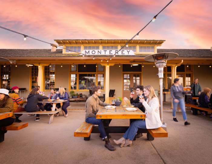 This is an image of the exterior of Dust Bowl Brewing Co. in Monterey at sunset. Various groups of friends sit at wooden bench style seating in the breweries outdoor area, laughing and drinking beer together.