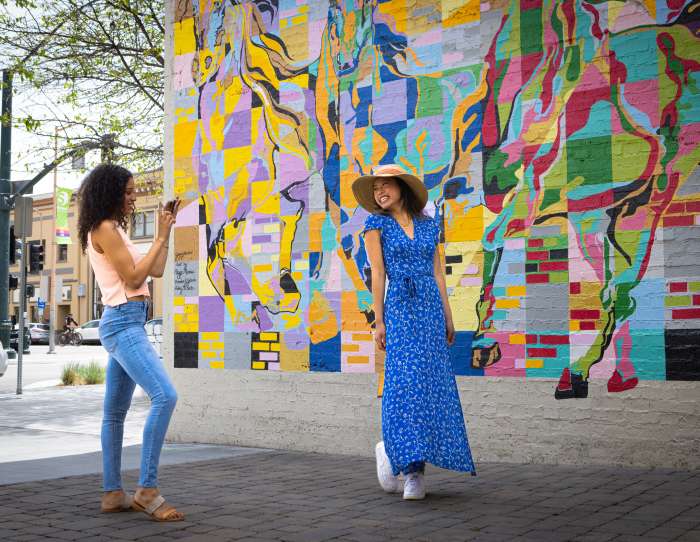 This is an image of two women in front of a colorful mural in Downtown Salinas. One woman is smiling and posing in front of the rainbow colored mural while the other women stands nearby taking a photo of her.