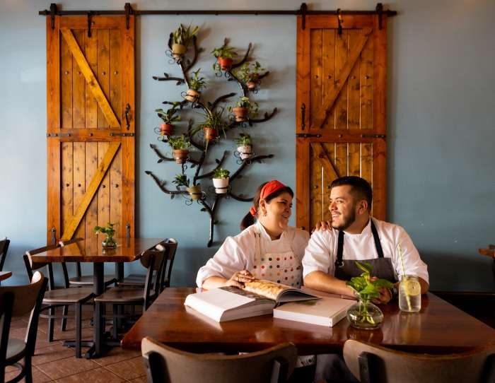 This is an image of the owners of Villa Azteca restaurant in Salinas. The owners are a mother and a son who are sitting next teach other on a table in the interior of their restaurant. The walls are blue sky and the decorations on the wall behind them include plants and a wooden farm door. The mother and son duo are looking at each other and smiling as they sit on of of the restaurants tables. There is a couple of books opened in front of them, a small vase of plants, and an ice cold water with a lemon. They both wear aprons and white chefs shirts.