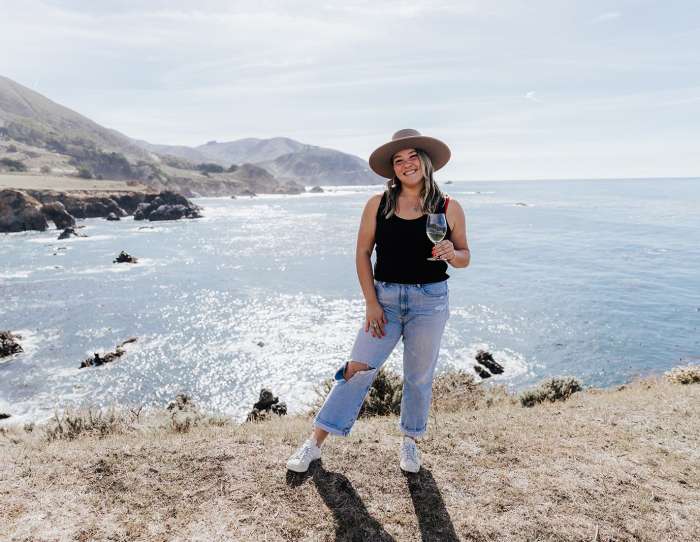 This is an image of Ashley Wolff, owner of Pangaea Grill and Jeju Kitchen in Carmel-by-the-Sea, standing in front of the Big Sur coastline holding a glass of wine