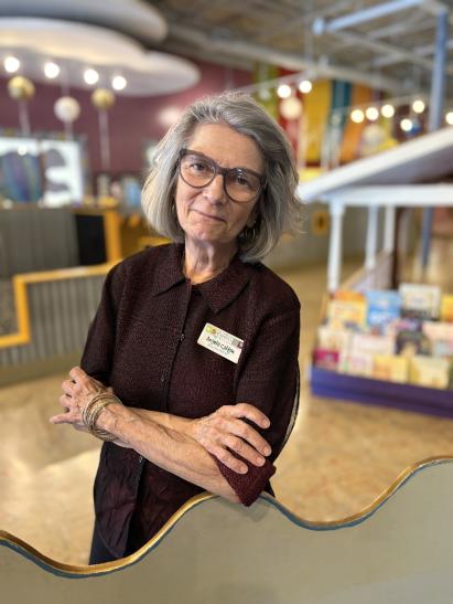 A headshot of a woman (60s) facing the camera. She has short silver hair and large glasses. She is wearing a textured maroon button-down shirt and a nametag with her name and the museum's. Her arms are crossed lightly over her chest with one elbow resting on a children's play structure shaped like an ocean wave. Behind her, though out of focus, are various colorful kid's museum displays.