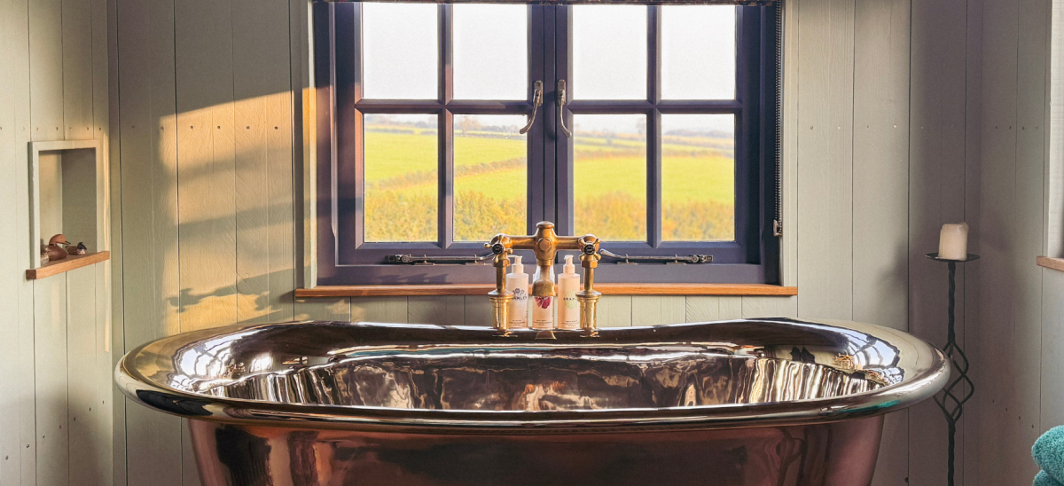 photo shows a plush metal bath tub in a shepherds hut, looking out through a window at countryside views