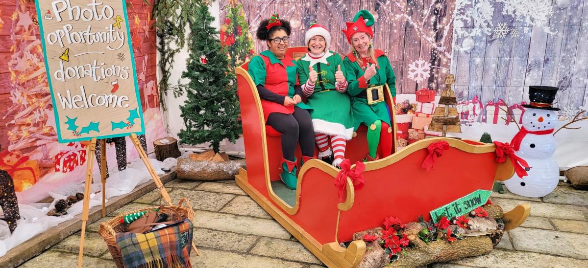 Three women dressed as Christmas characters sitting in a red sleigh