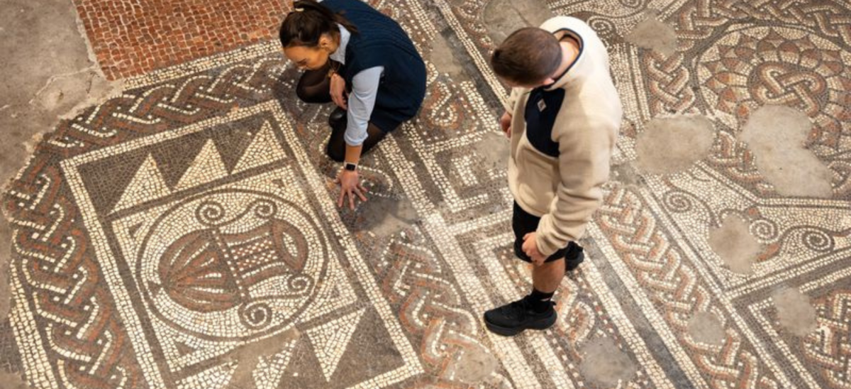 An aerial shot looking down on two men looking at the roman floor mosaic at Dorset museum. One, the guide, is kneeling on the floor and explaining.