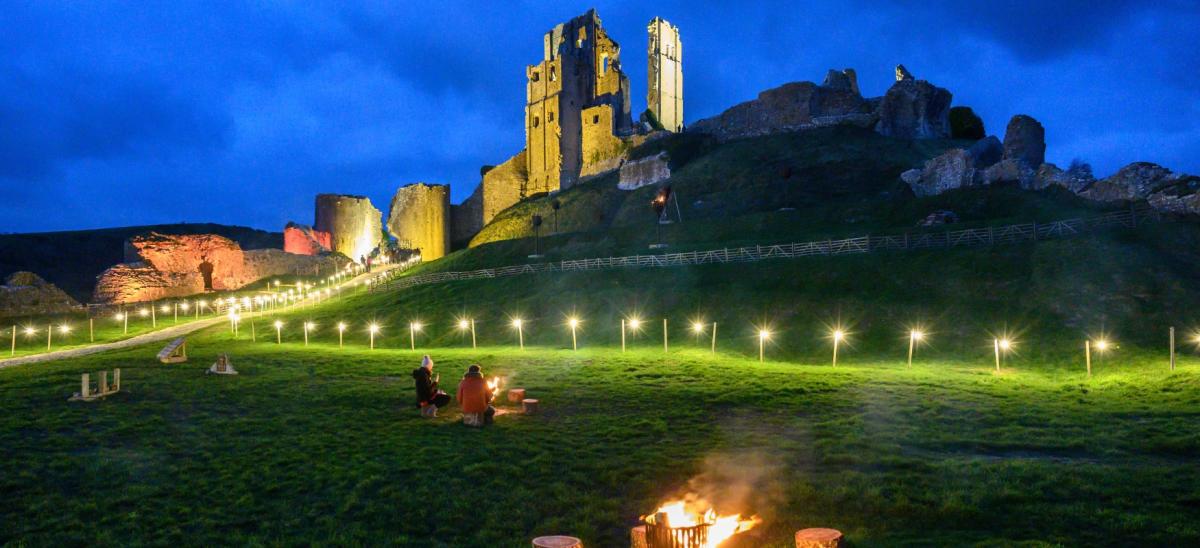 Two people toasting marshmallows looking towards an illuminated Corfe Castle during winter