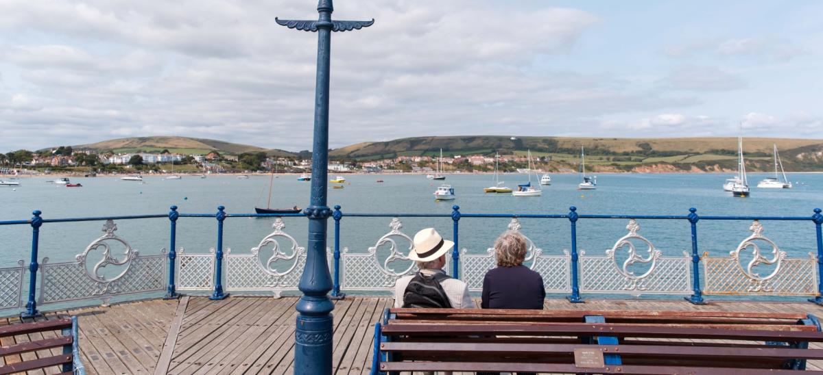 Two people sat on a bench on Swanage Pier in Dorset looking towards the Purbeck hills