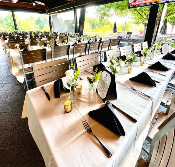 Multiple tables are set for a group or private dining event with white table linens and black napkins.. The windows overlook some green.