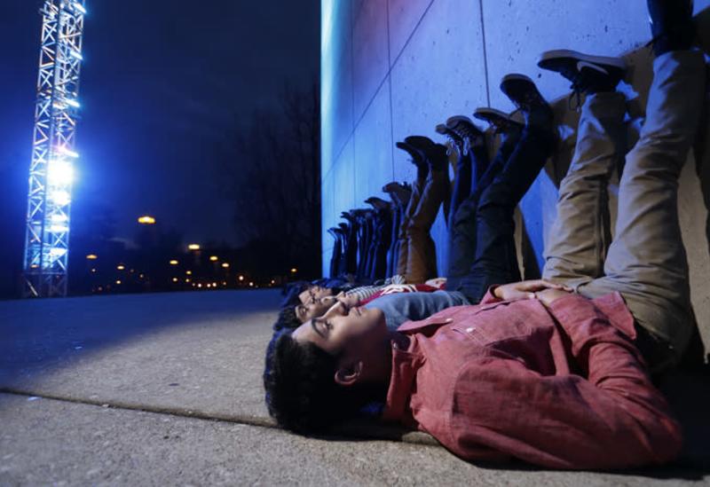 Students laying on the ground at night with their legs propped up against the IU Art Museum as the Light Totem colors change