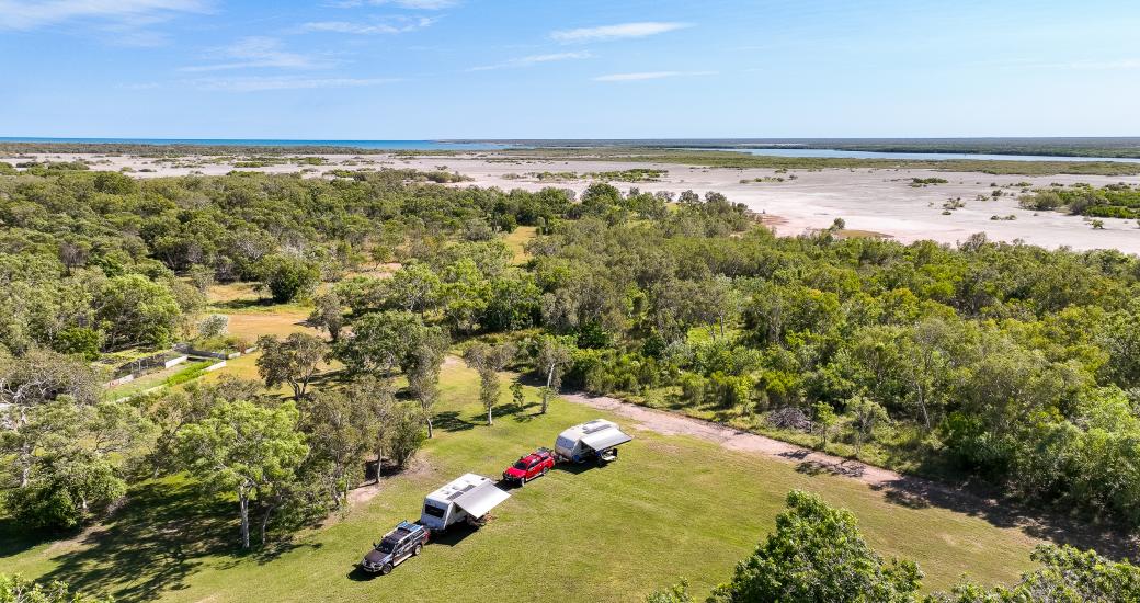 Aerial view of cars and caravans in the grassed campground at Banana Well Getaway, with the ocean visible in the background