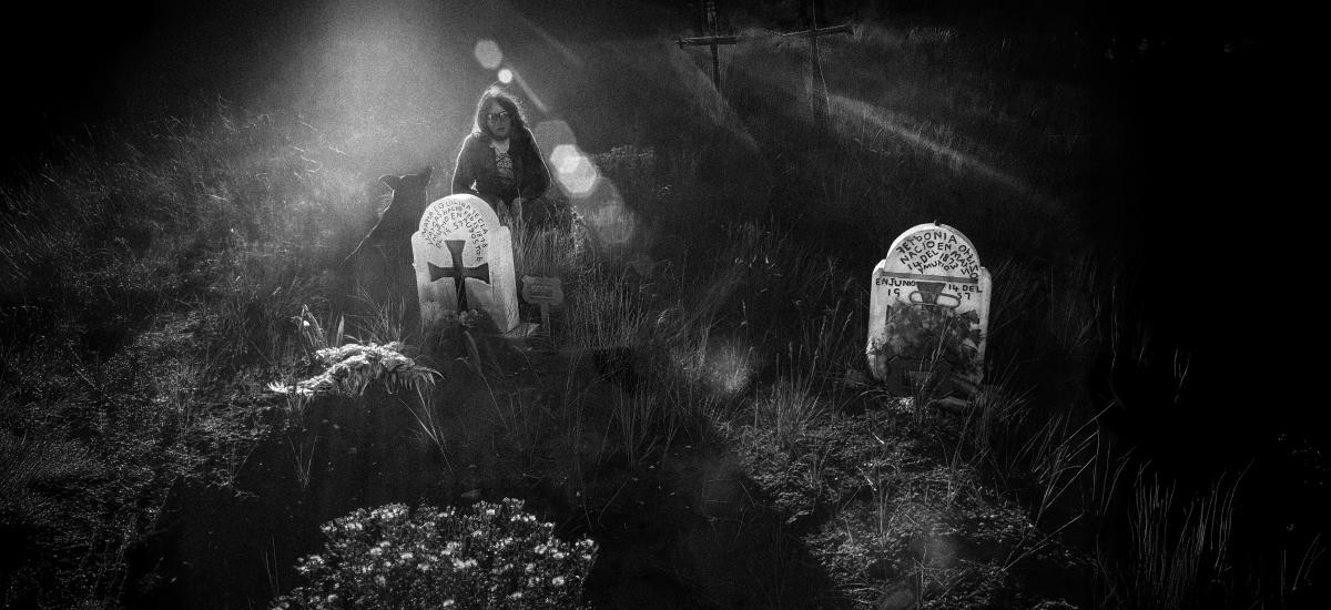 Black and white photograph of a woman standing among weathered gravestones in a grassy cemetery.
