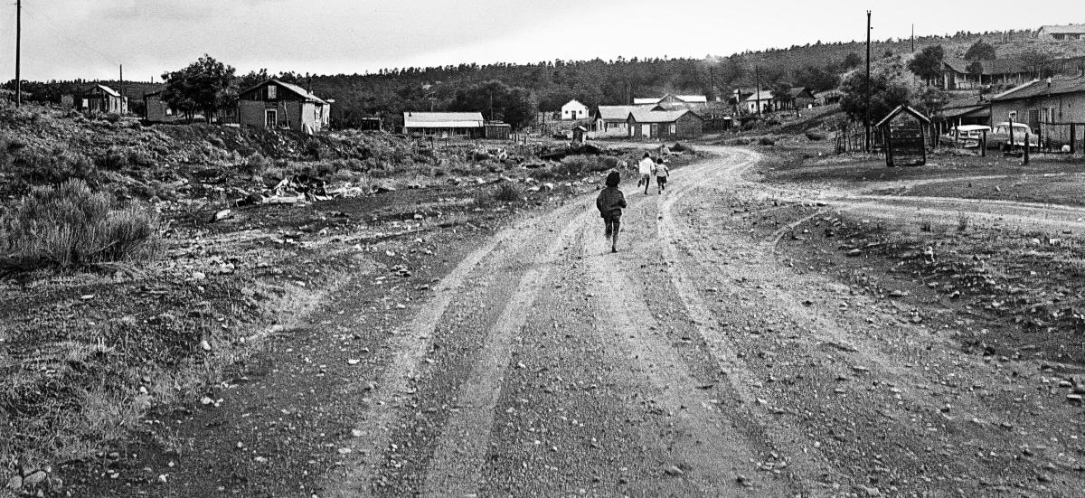 Black and white landscape photograph of several children walking along a wide dirt road through the village of Petaca.