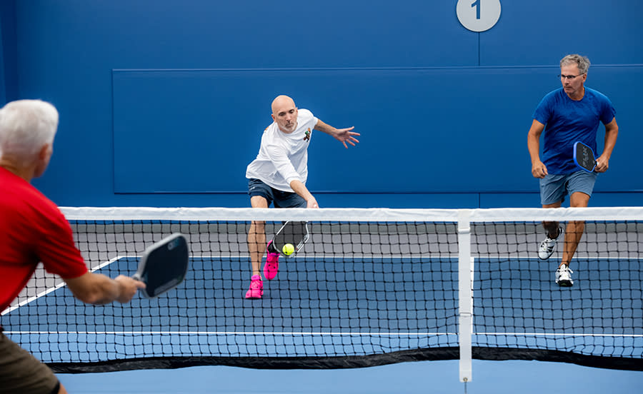 a group of three men playing pickleball, one running up to the net to return the ball